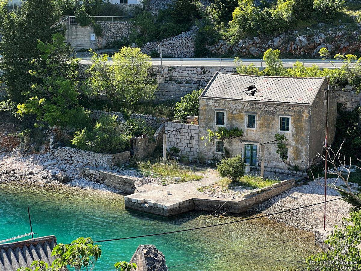 Altes, renovierungsbedürftiges Steinhaus direkt am Wasser mit beschädigtem Dach und umgebender Natur.