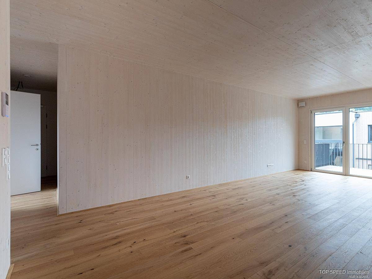 Geräumiger, unmöblierter Wohnbereich mit Holzboden und Zugang zu einem Balkon.
