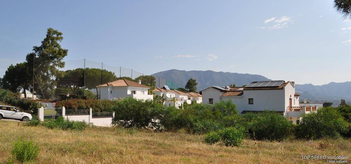 Weitläufige Landschaft mit Wohnhäusern, Tennisplatz und Bergen im Hintergrund unter blauem Himmel.