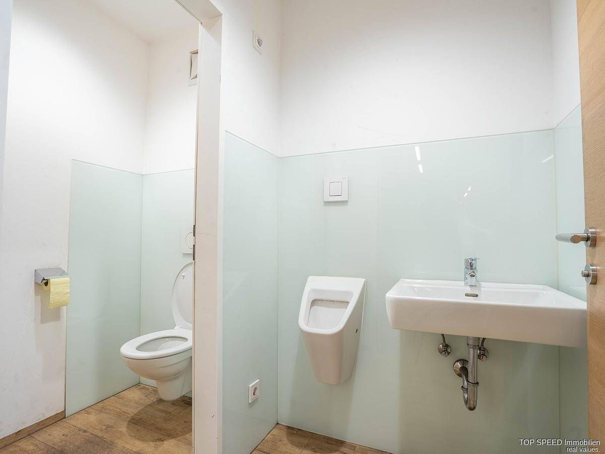 Modernes Badezimmer mit Toilette, Urinal und Waschbecken, ausgestattet mit hellen Fliesen und Holzboden.