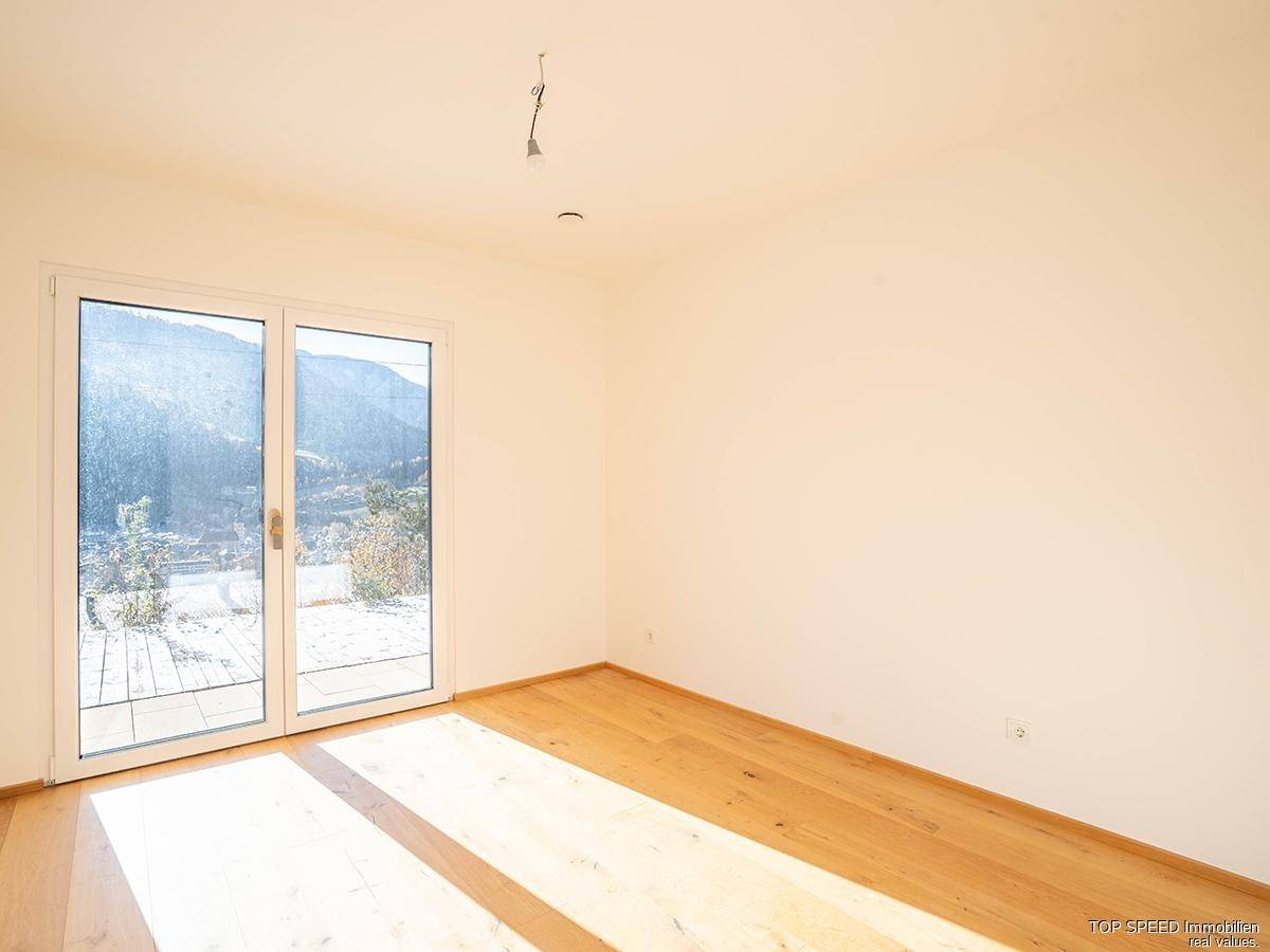 Helles, leeres Zimmer mit Holzboden und großen Fenstern, die viel Sonnenlicht hereinlassen.