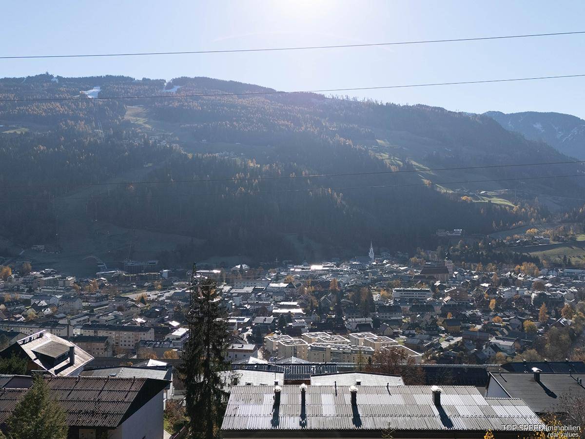 Weitläufiger Blick über eine Stadt im Tal, umgeben von bewaldeten Hügeln unter blauem Himmel.