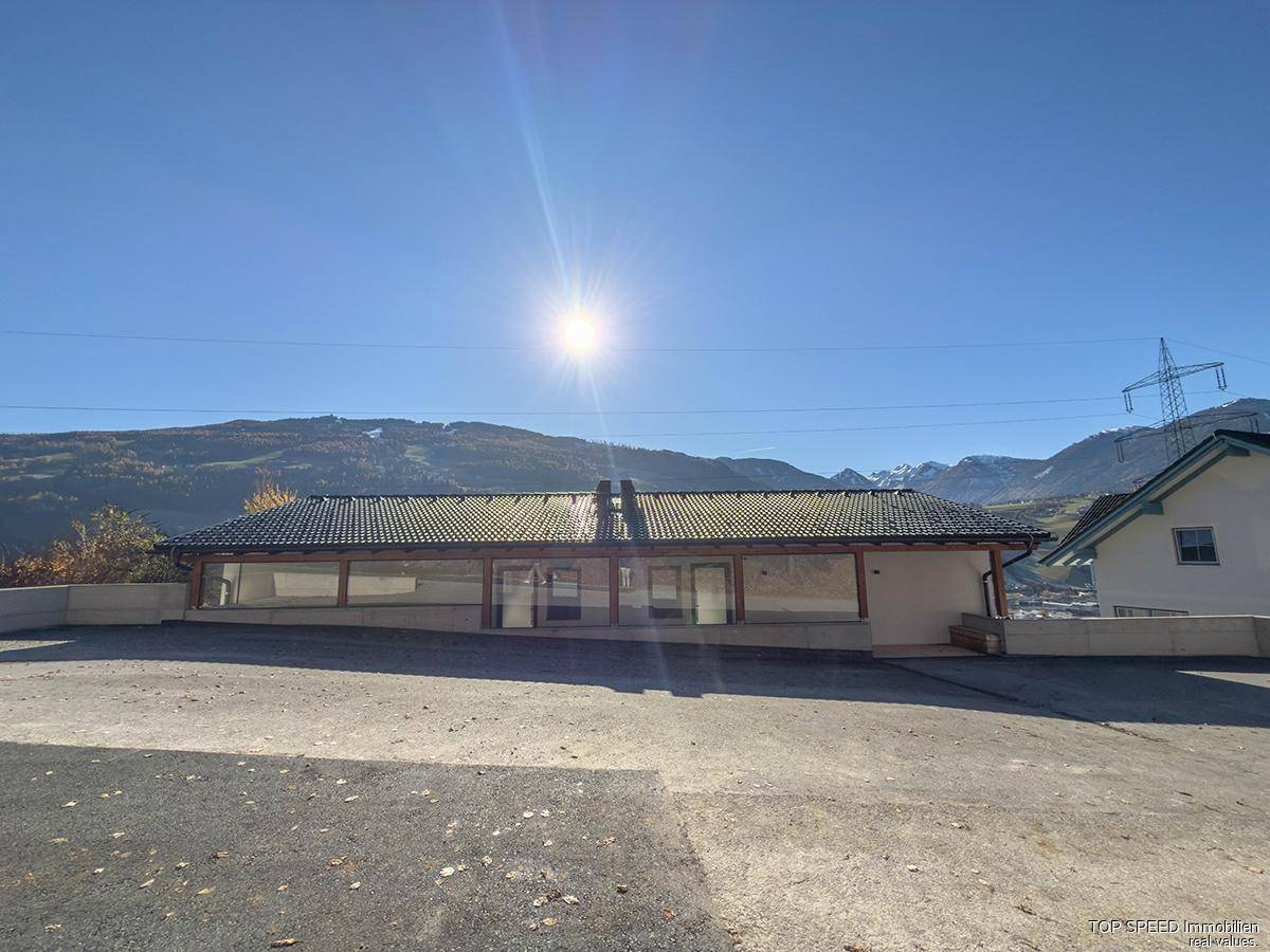 Neubau mit großem Vorplatz und Satteldach, umgeben von Bergen unter strahlendem Sonnenschein.