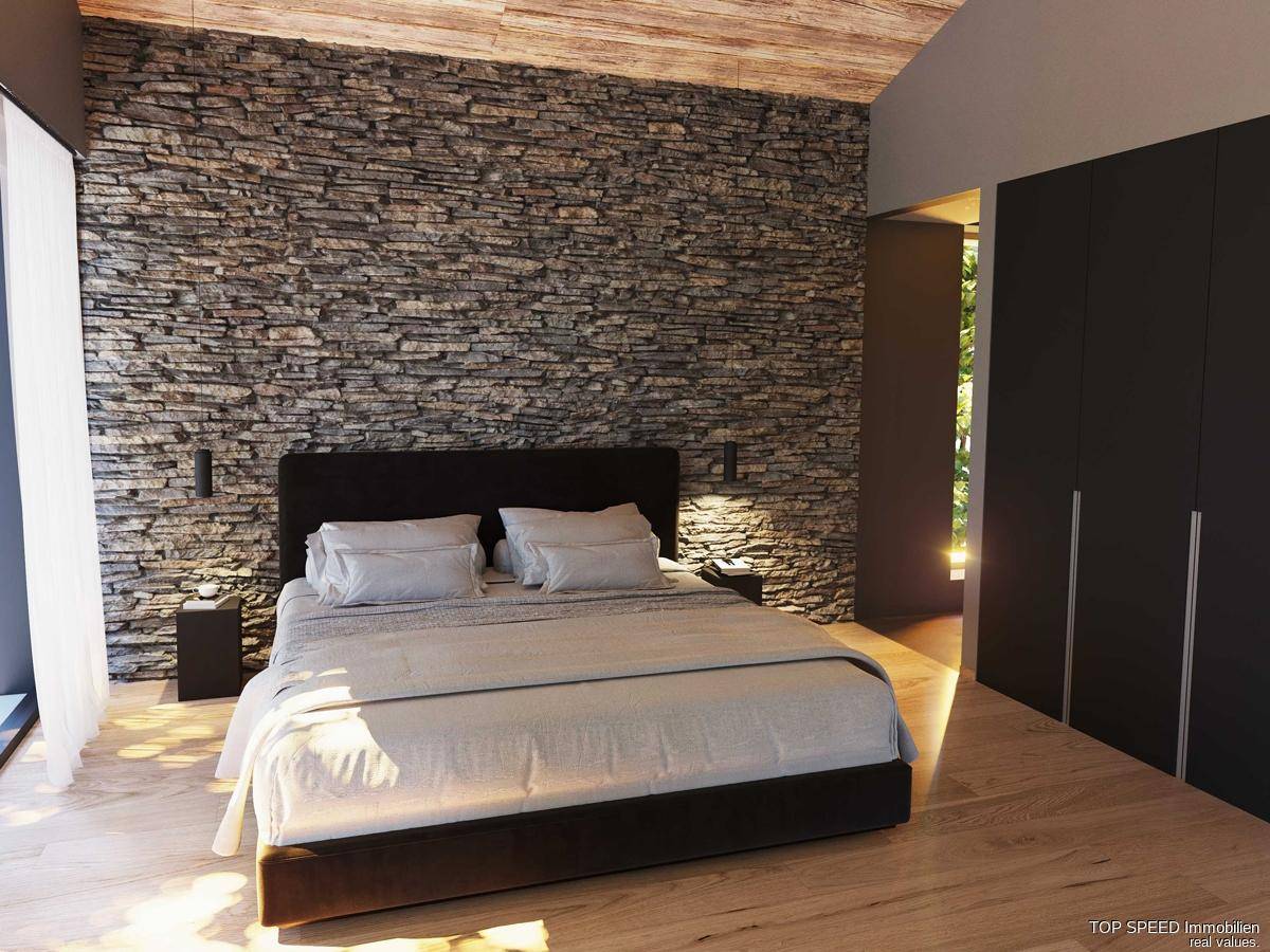Schlafzimmer mit markanter Steinwand, Holzbalkendecke und großem Bett, hell beleuchtet durch Fenster.