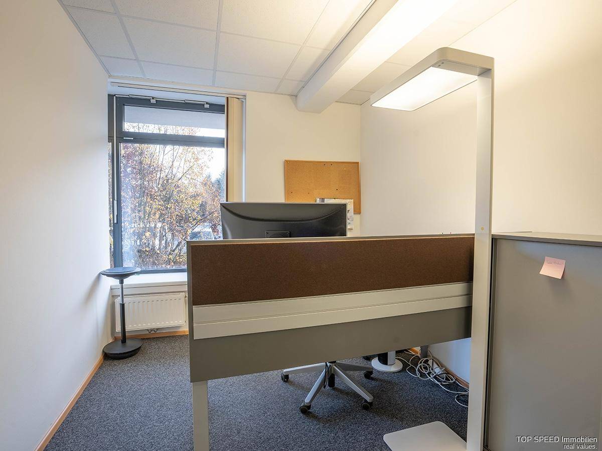 Einzelner Arbeitsplatz in einem Büro mit Schreibtisch, Monitor und Fenster mit Blick nach draußen.
