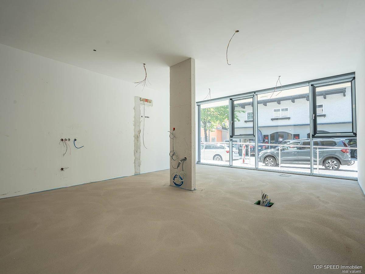Unfertiger Innenraum mit großen Fenstern zur Straße und sichtbaren Installationen für den Ausbau.