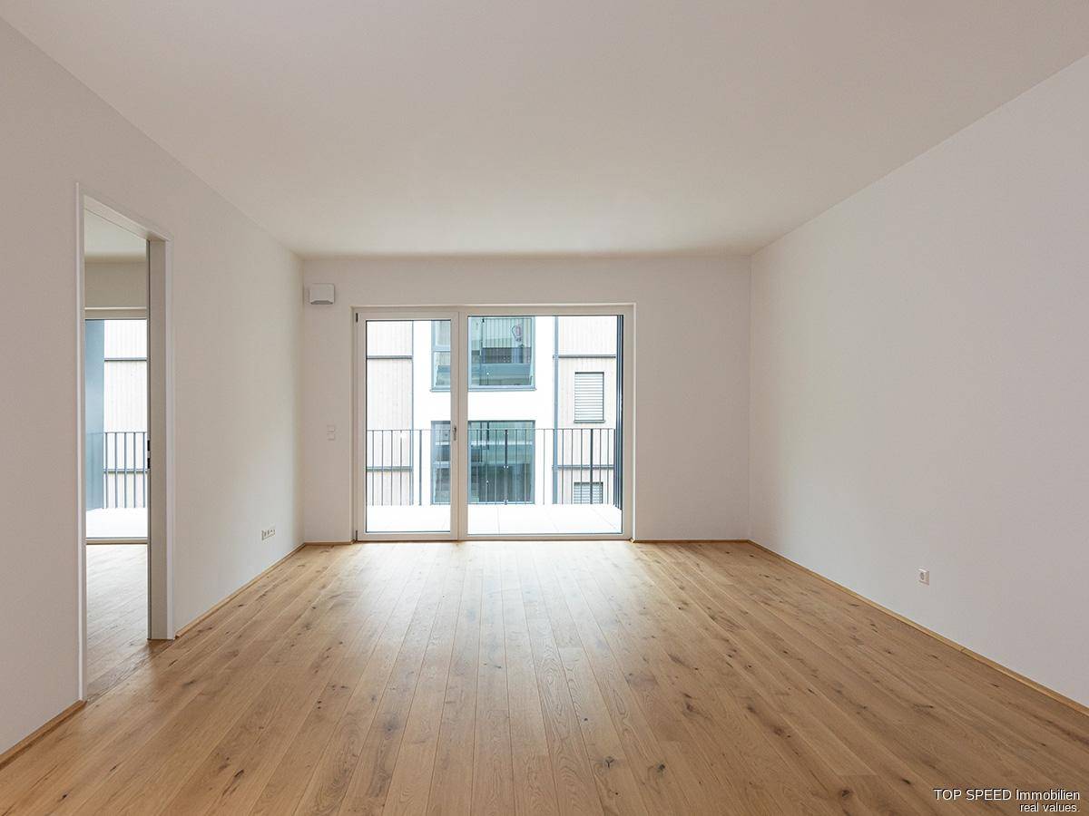 Geräumiger Wohnbereich mit Holzboden und breiter Balkontür, die viel natürliches Licht hereinlässt.