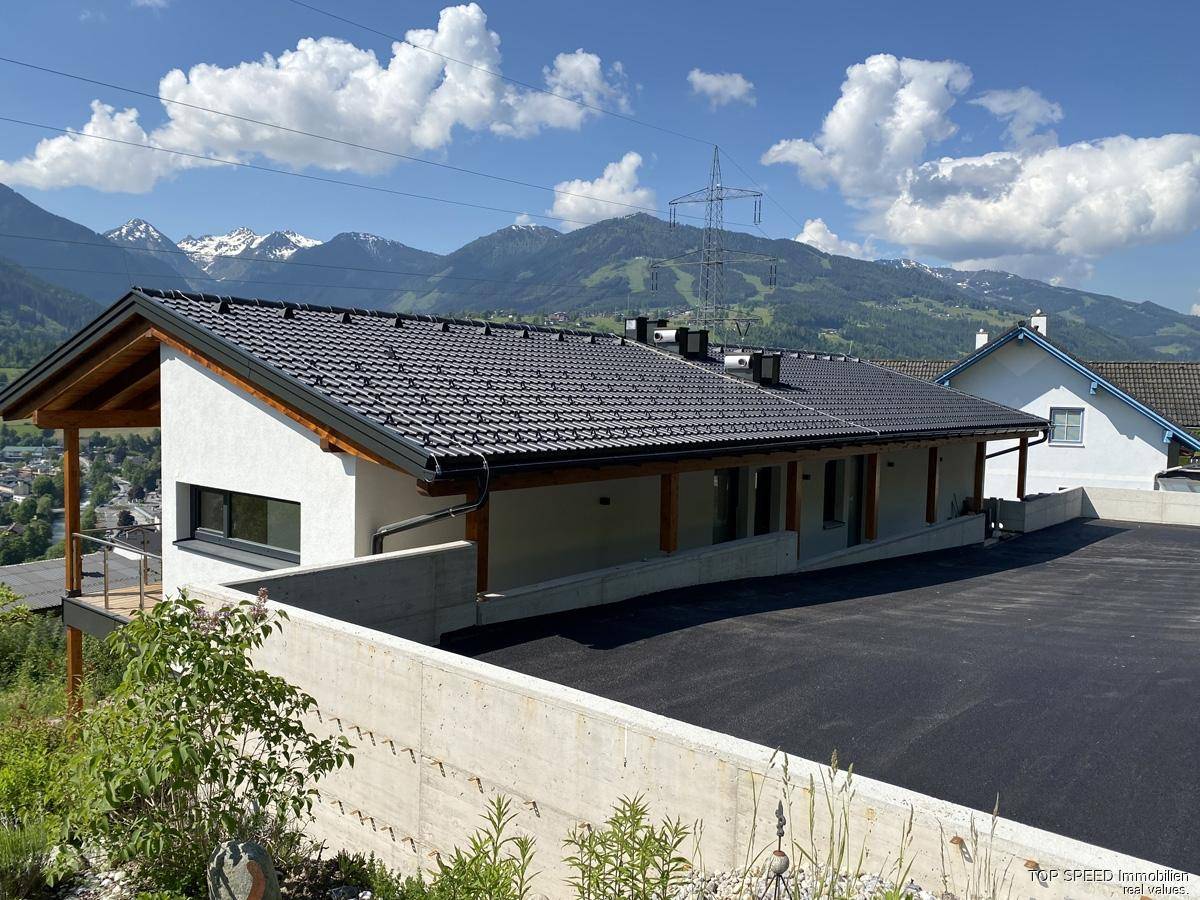 Modernes Gebäude mit Satteldach und großer Auffahrt, umgeben von Bergen und grüner Landschaft.