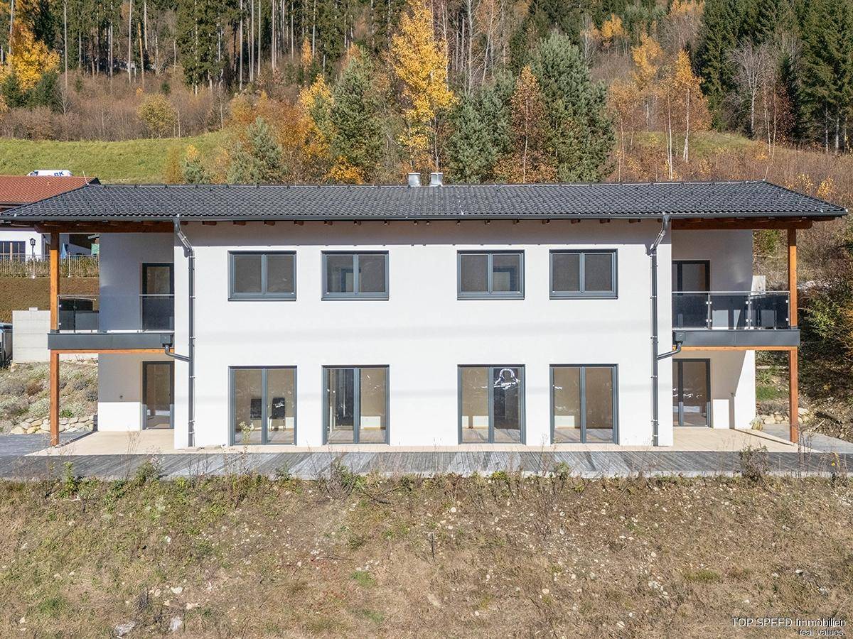 Moderne Gebäudefassade mit weißen Wänden, dunklen Fenstern und Balkonen vor einem bewaldeten Hang.
