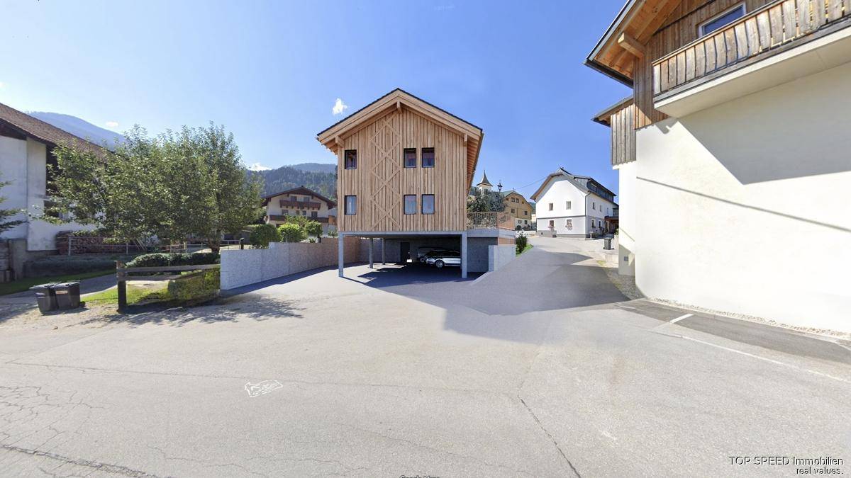 Modernes Holzgebäude auf Stelzen mit überdachtem Parkplatz und Bergblick, eingebettet in die Landschaft.