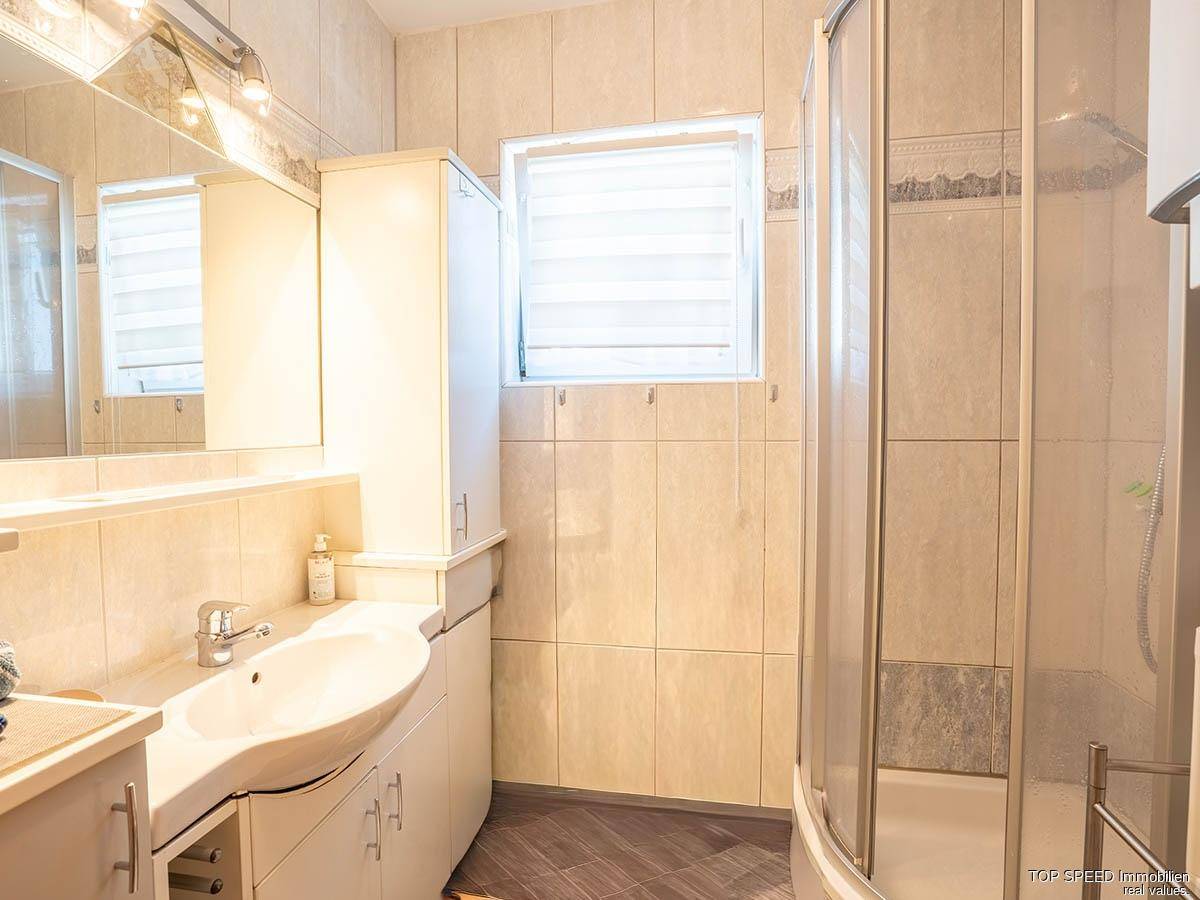 Gefliestes Badezimmer mit Waschbecken, Spiegelschrank und einer Duschkabine, ausgestattet mit einem Fenster.