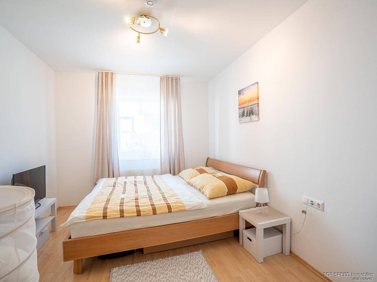 Helles Schlafzimmer mit einem Holzbett, Nachttisch und einem Fenster, das viel Tageslicht hereinlässt.
