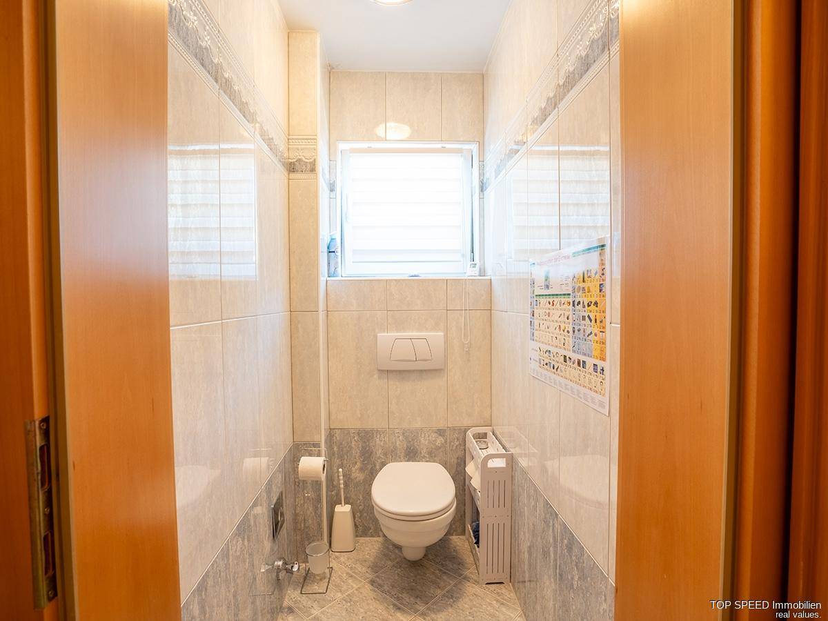 Separates WC mit hellen Fliesen, wandhängendem WC und einem Fenster für natürliche Belüftung.