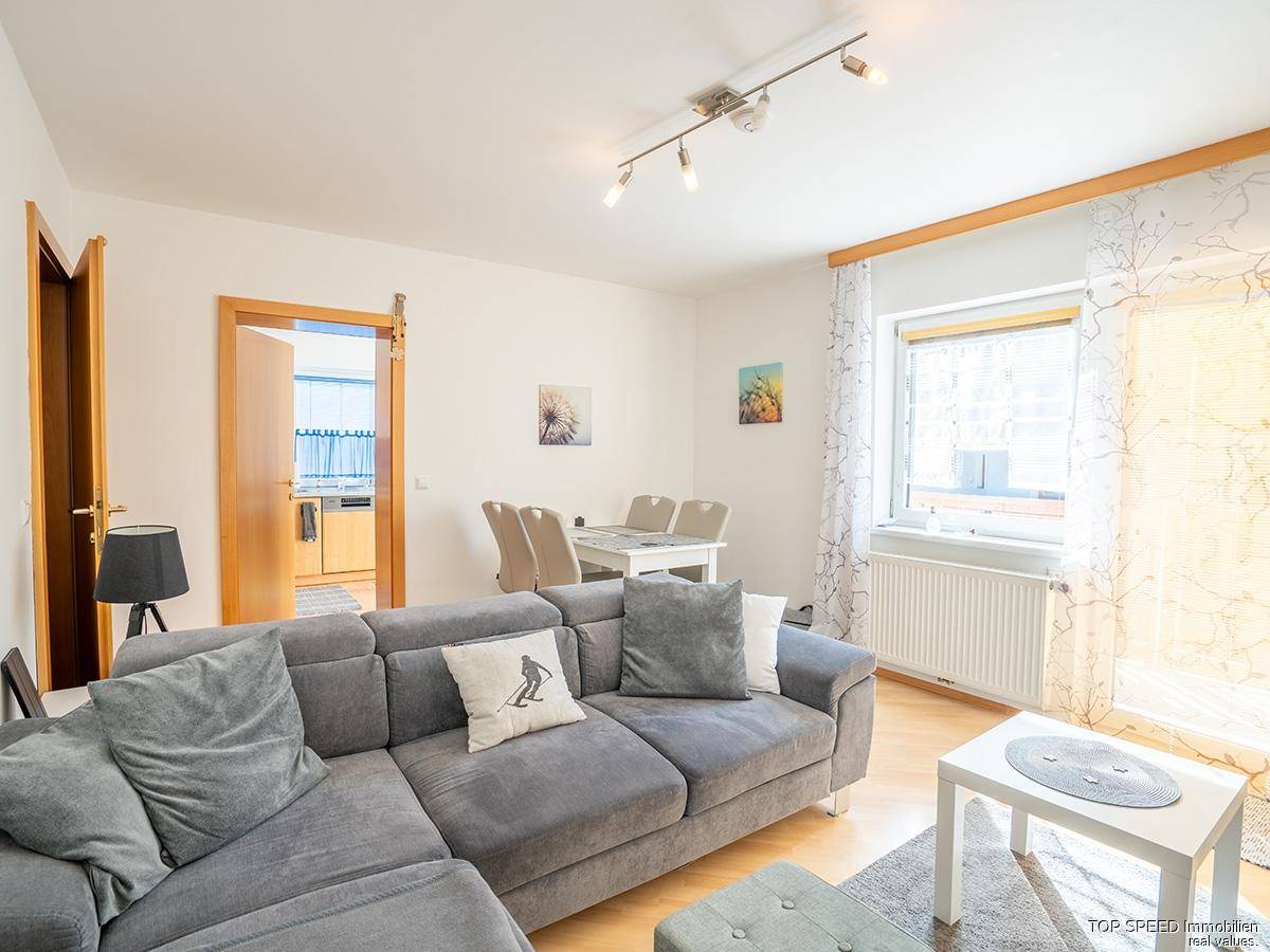 Gemütlicher Wohn-Essbereich mit grauem Ecksofa, hellem Holzboden und einem Esstisch im Hintergrund.