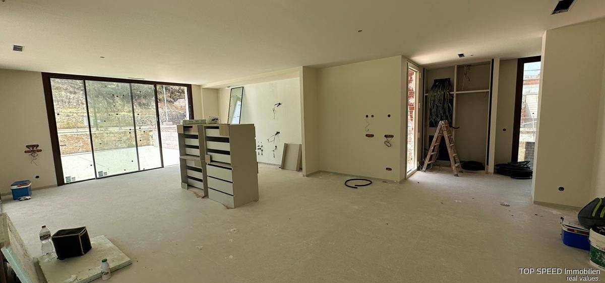 Großer Innenraum im Rohbau mit großen Fensterfronten und Blick ins Freie.