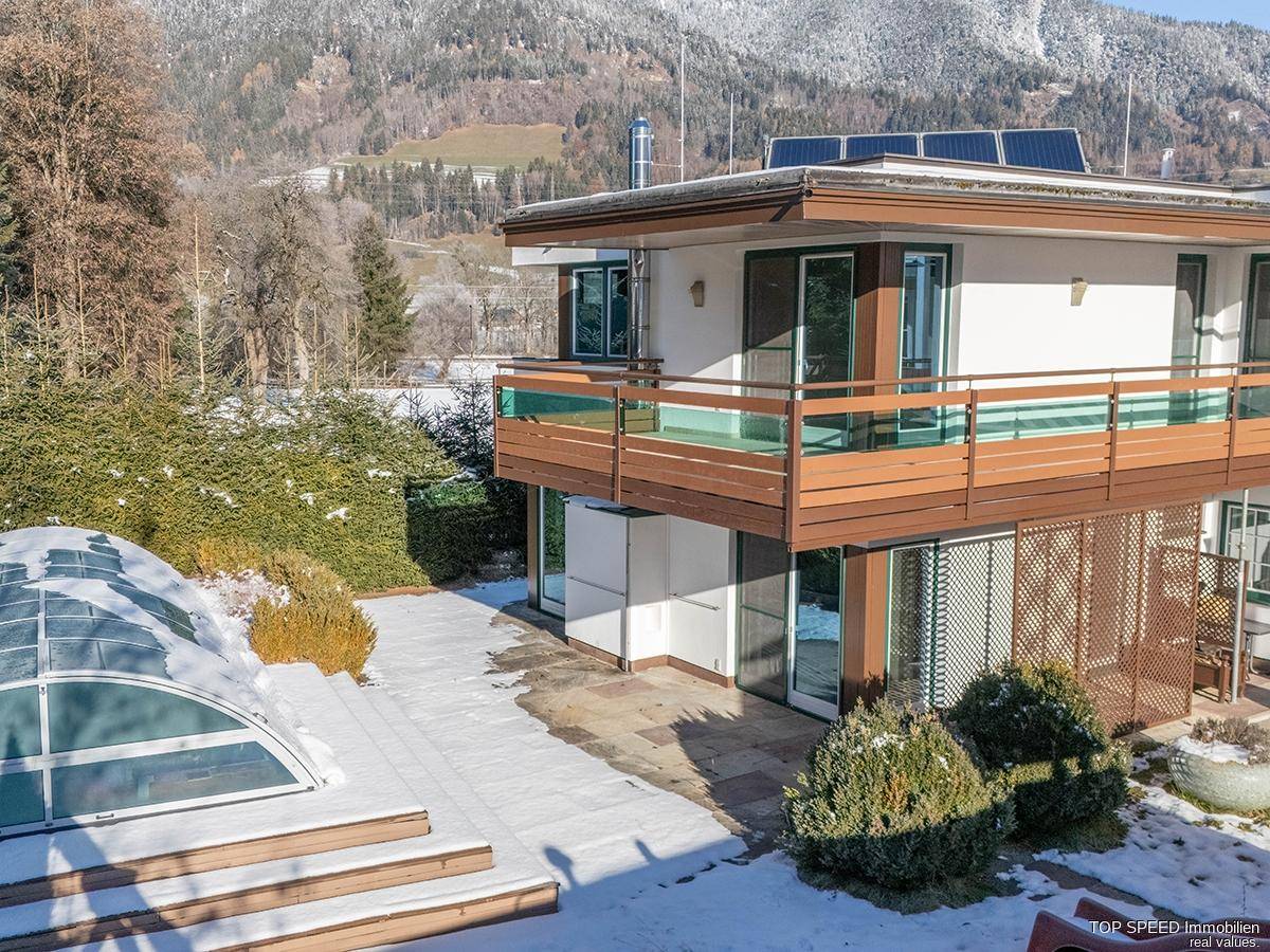 Ansicht des Hauses mit Balkonen, Solarpaneelen und überdachtem Pool vor verschneiter Bergkulisse.