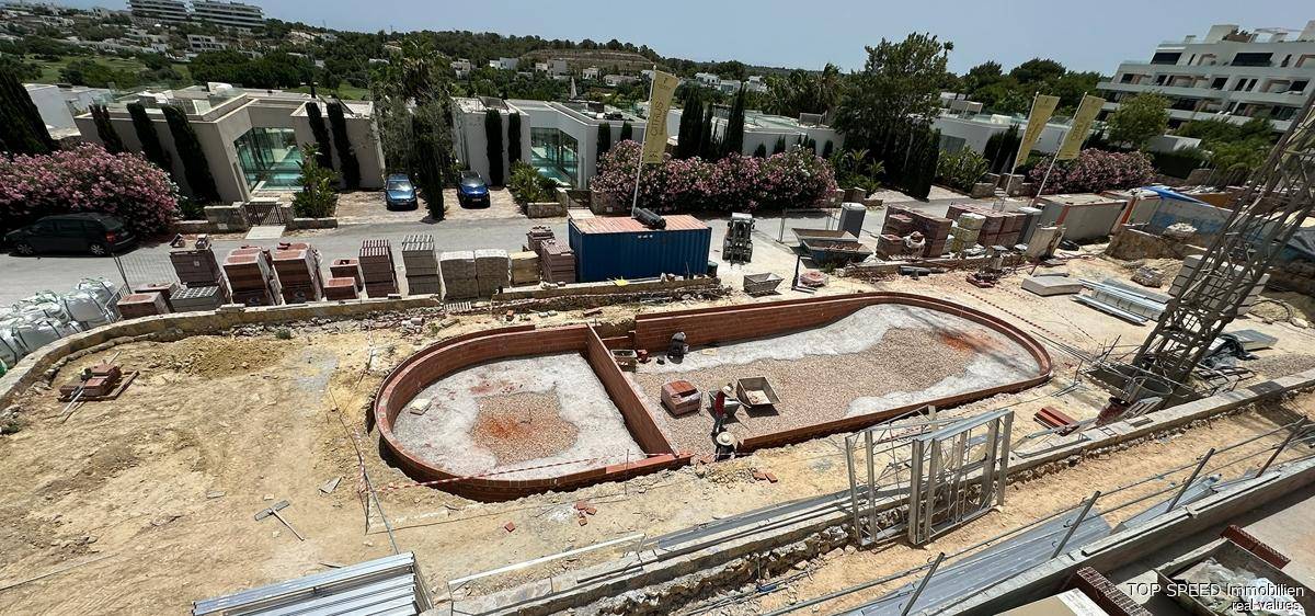 Luftaufnahme einer Baustelle mit Poolfundament und Baumaterialien, umgeben von Gebäuden.