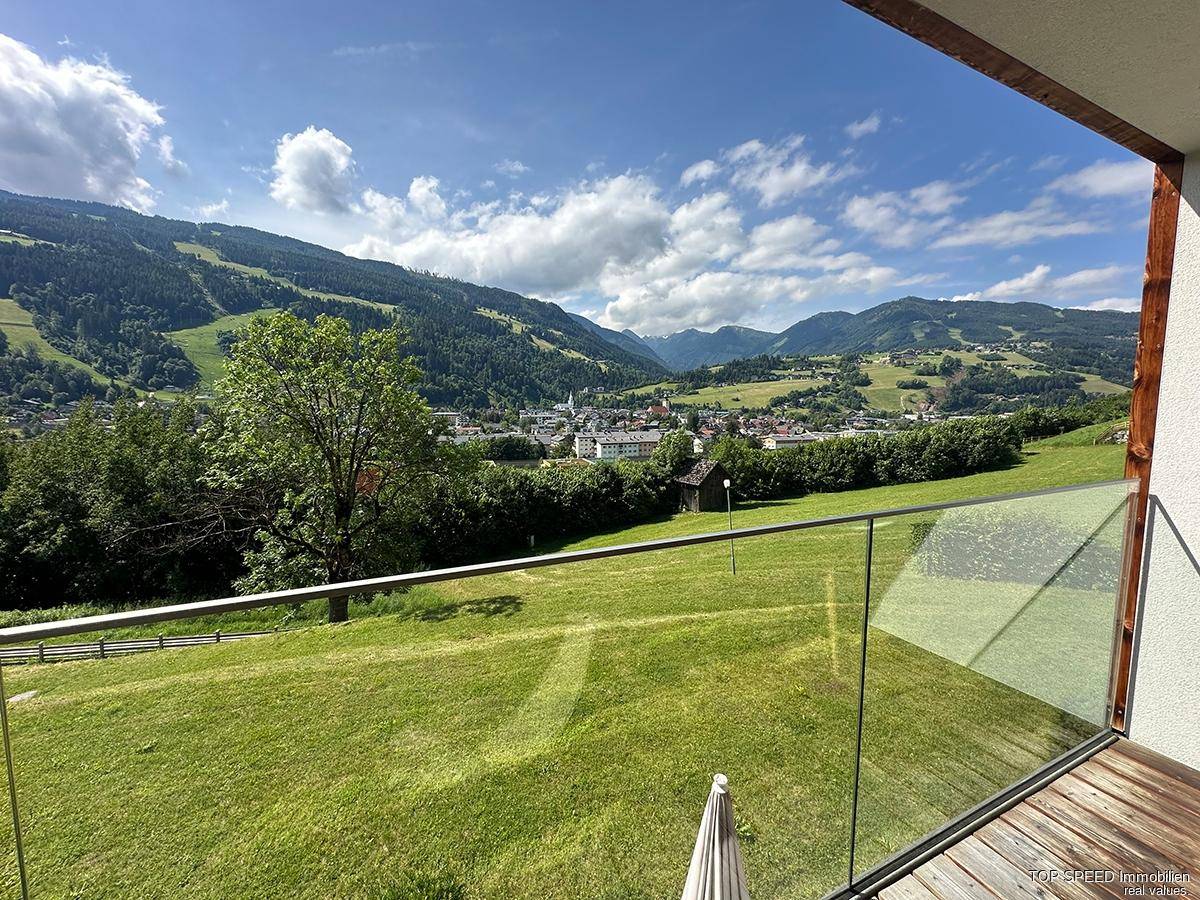 Weitläufiger Blick von einem Balkon über eine grüne Landschaft, ein Dorf und die umliegenden Berge.