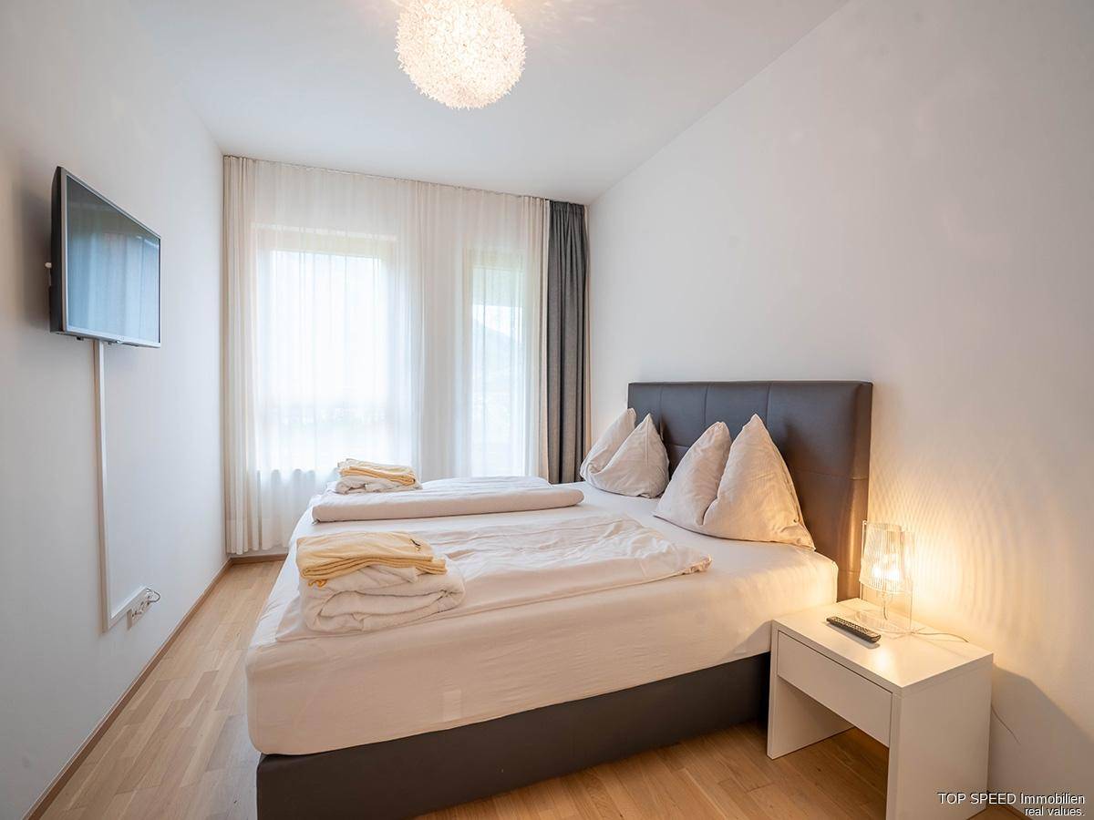 Komfortables Schlafzimmer mit Doppelbett, Wandfernseher und Fenster für angenehmes Licht.
