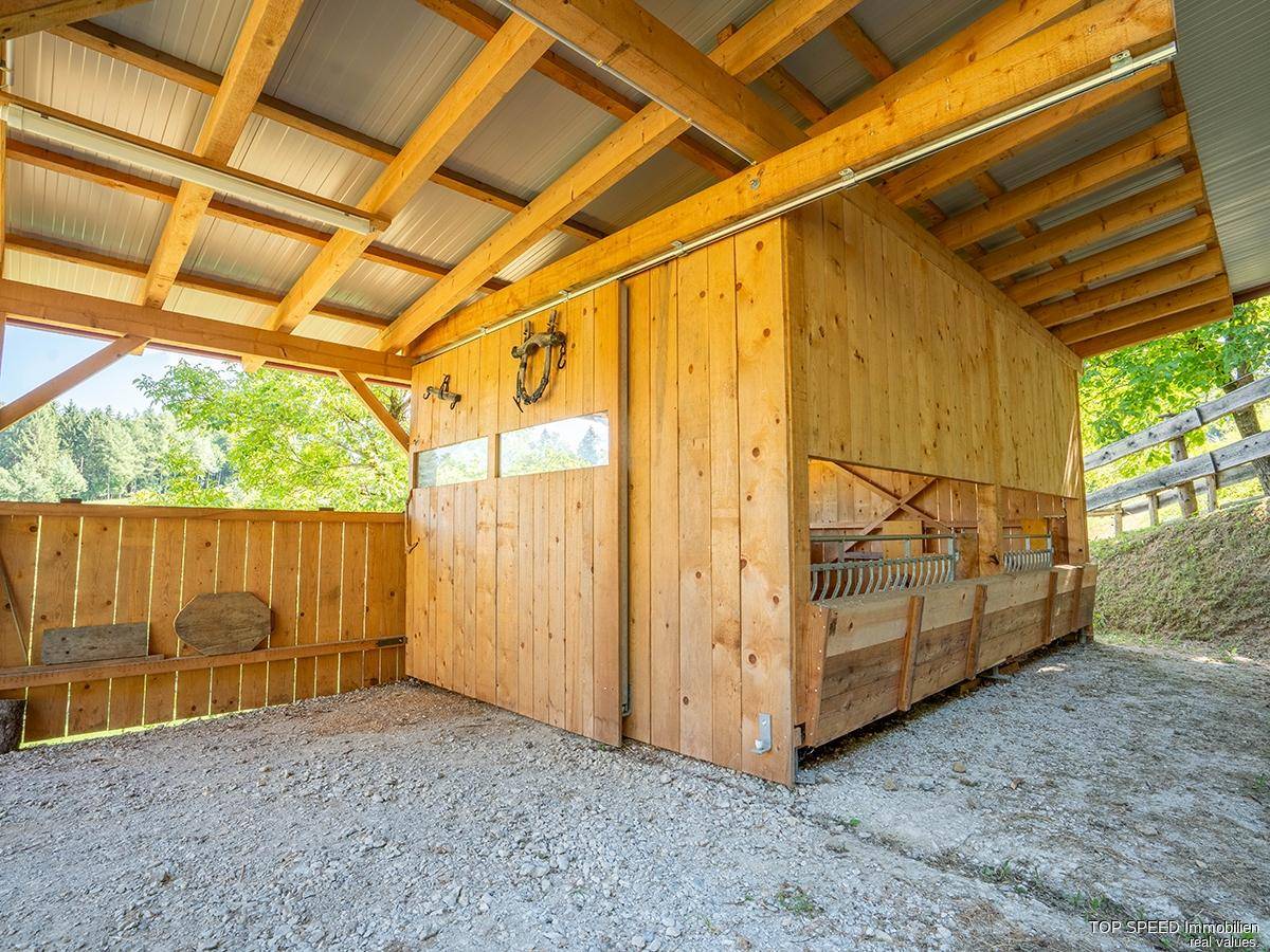 Offener Holzstall mit Boxen und Holzbalken, umgeben von grüner Natur, ideal für Tierhaltung.