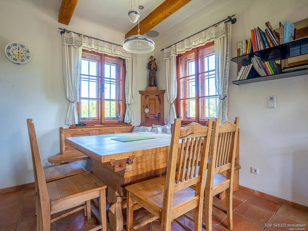 Traditioneller Essbereich mit Holztisch und Stühlen, sichtbaren Deckenbalken und Fenstern mit Blick ins Grüne.