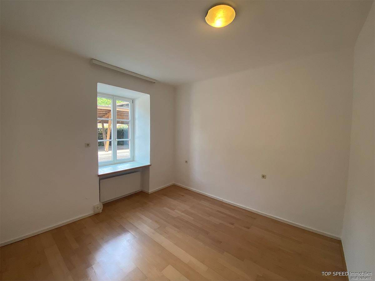 Leeres Zimmer mit Parkettboden und einem Fenster, das viel Tageslicht hereinlässt.