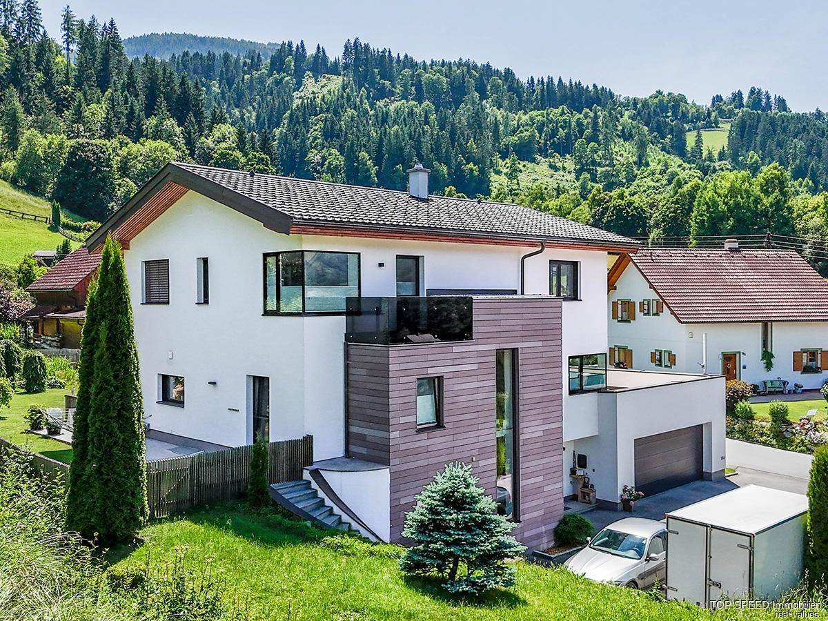 Modernes Wohnhaus mit integrierter Garage, Balkon und Blick auf die bewaldeten Berghänge.