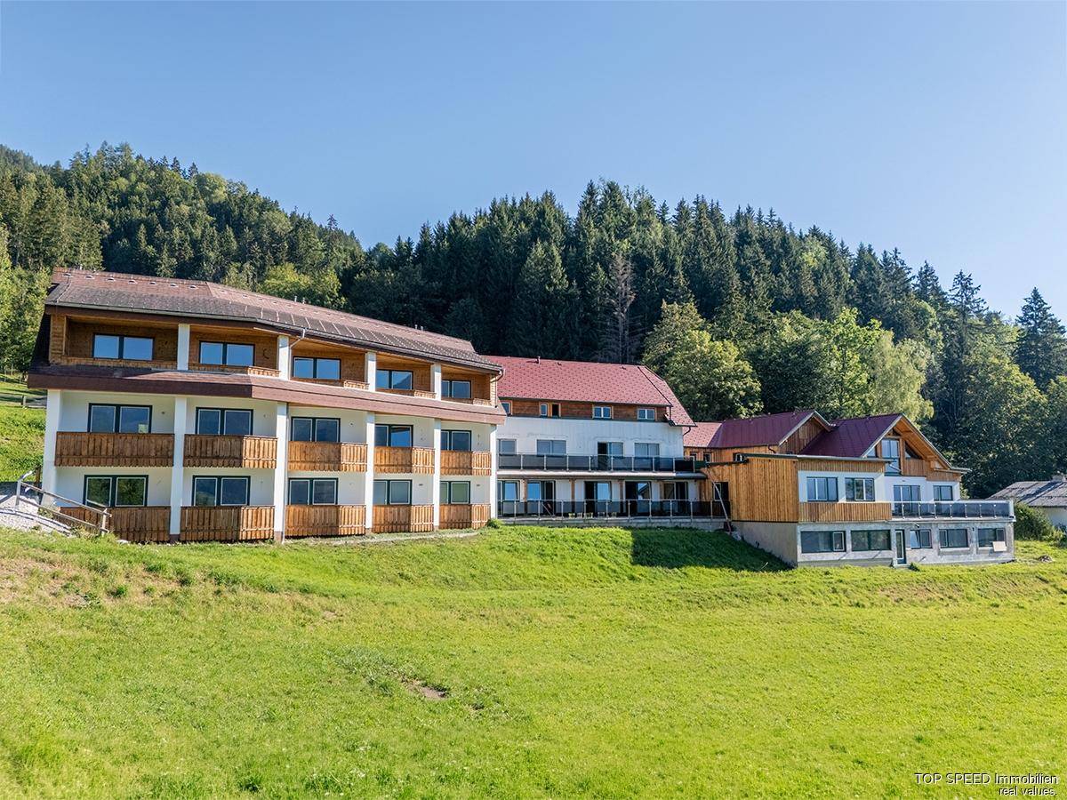 Mehrstöckiges Gebäude mit Balkonen, umgeben von grünen Wiesen und einem Wald im Hintergrund.