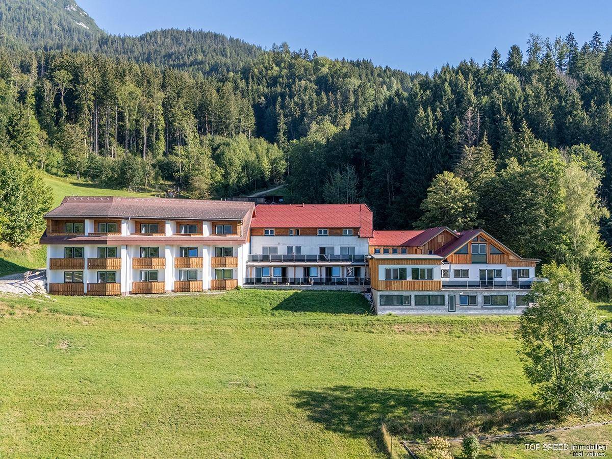 Weitläufige Ansicht eines modernen Gebäudekomplexes in alpiner Landschaft mit Wald und Wiesen.