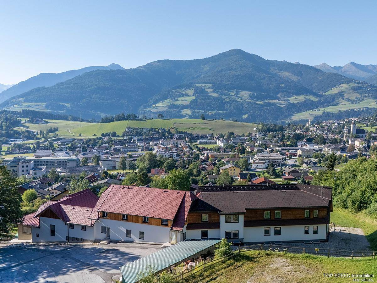 Luftaufnahme eines Gebäudekomplexes mit roten Dächern und einem Parkplatz, mit Blick auf eine Stadt und Berge im Hintergrund.