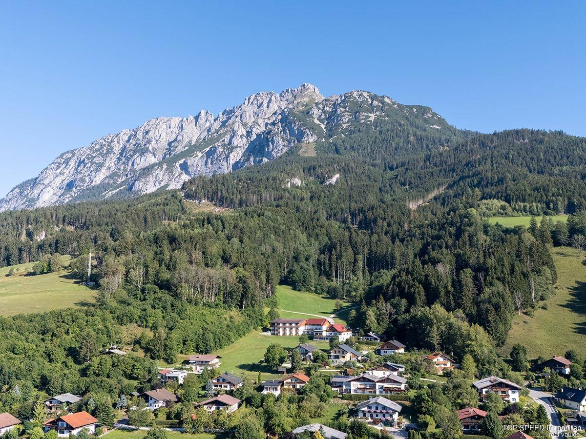 Weitläufige Luftaufnahme einer Siedlung am Fuße eines imposanten Berges, umgeben von dichten Wäldern und grünen Hängen.