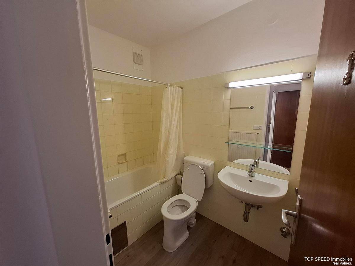 Funktionales Badezimmer mit Badewanne, WC, Waschbecken und hellbeigen Fliesen im 80er-Jahre-Stil.