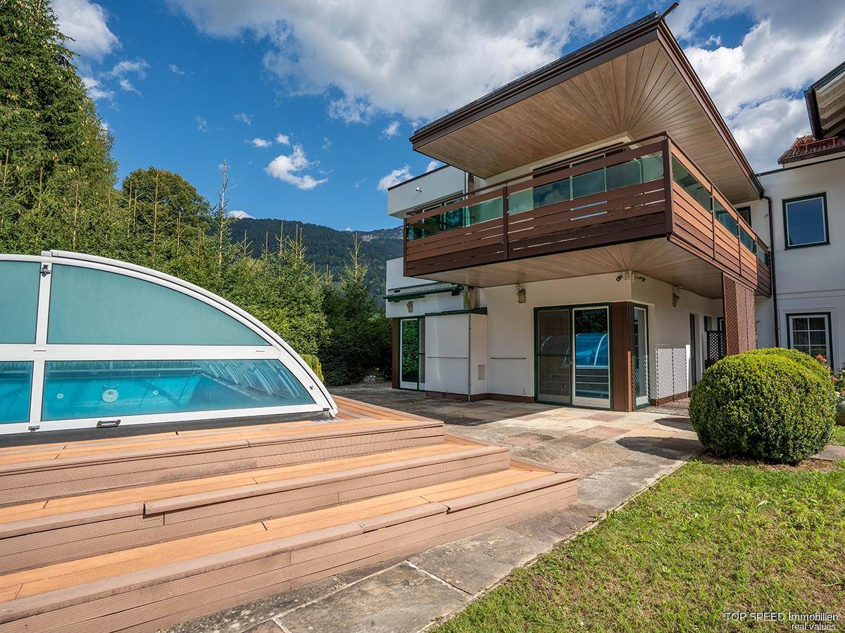 Hausfassade mit überdachtem Pool und Holzdeck, umgeben von viel Grün und Bergblick.