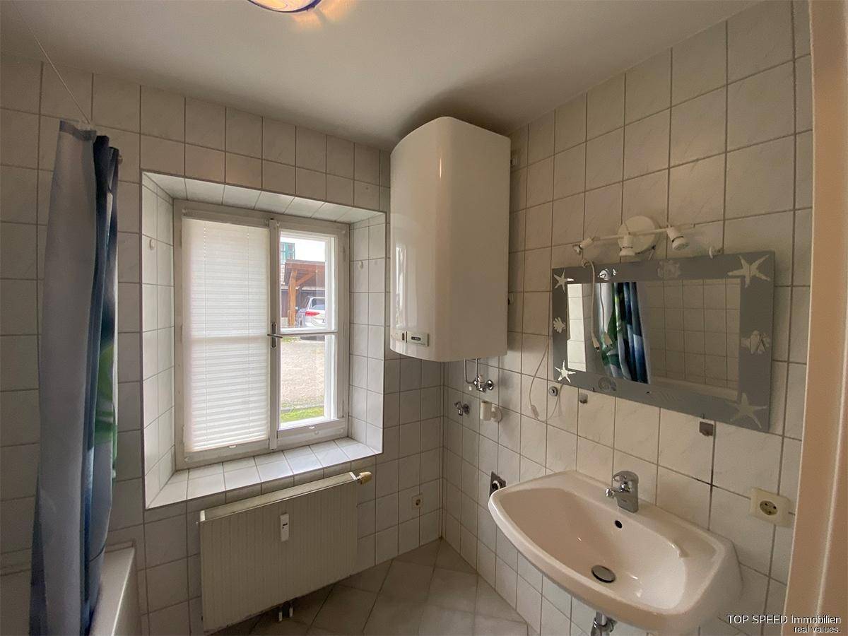 Gefliestes Badezimmer mit Waschbecken, Spiegel, Warmwasserboiler und Fenster mit Jalousie.