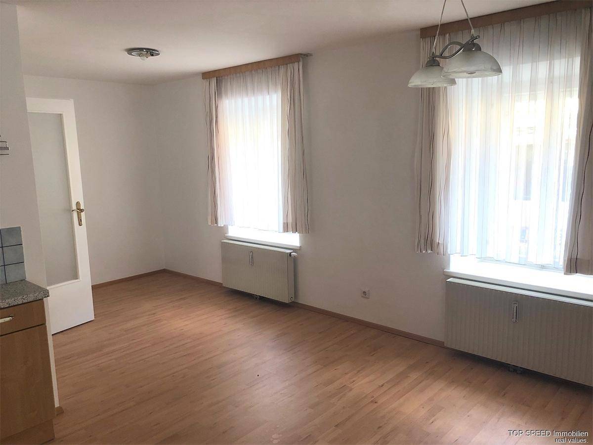 Heller Wohnraum mit Laminatboden, weißen Wänden und zwei Fenstern mit Vorhängen.