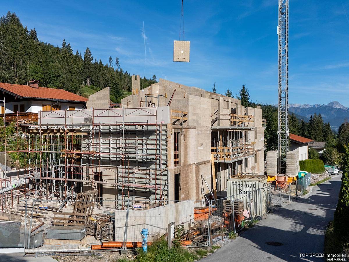 Baustelle eines neuen Wohngebäudes in den Bergen mit Gerüst und Kran unter blauem Himmel.