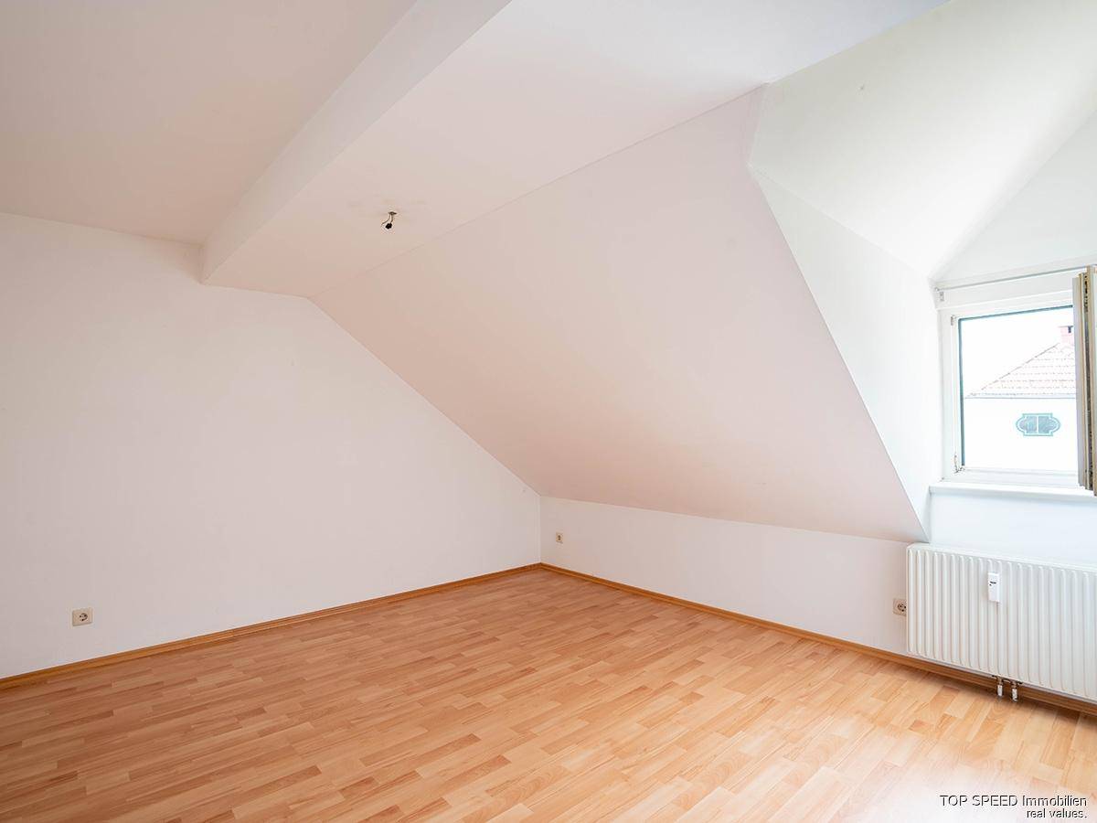 Leeres Zimmer im Dachgeschoss mit schrägen Wänden, Laminatboden und einem Fenster.