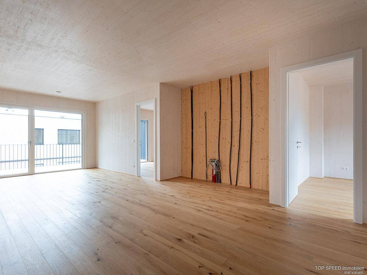 Heller, leerer Wohnraum mit Holzboden und Holzverkleidung, großen Schiebetüren zum Balkon und sichtbaren Installationen.