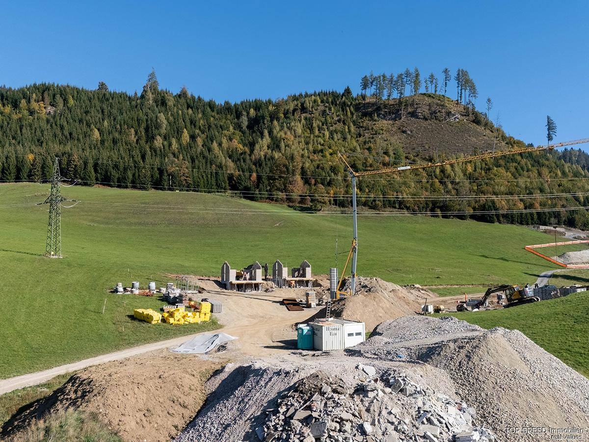 Weitläufige Baustelle mit Kran und mehreren im Bau befindlichen Gebäuden vor bewaldeten Bergen.
