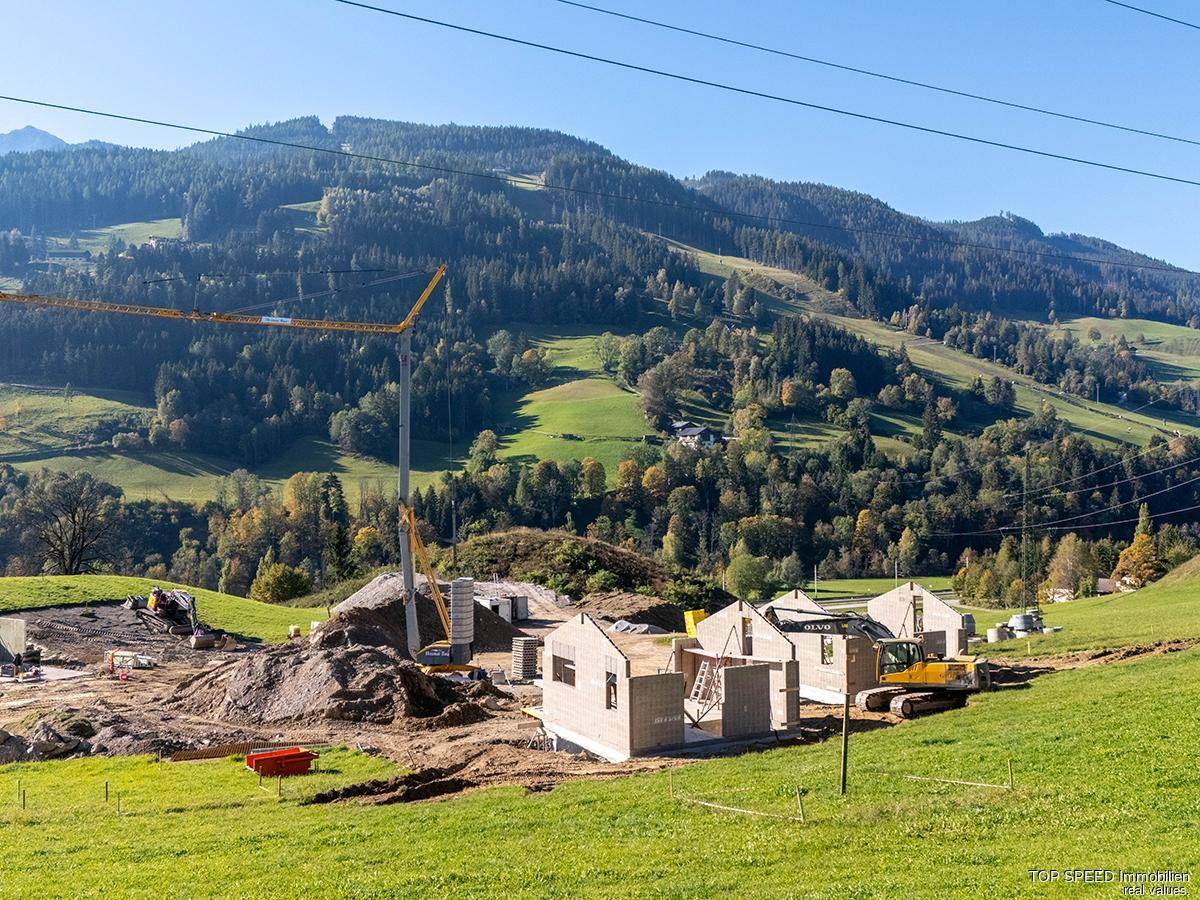 Baustelle mit mehreren Rohbauten und Baugeräten, eingebettet in eine malerische Landschaft mit grünen Hügeln.