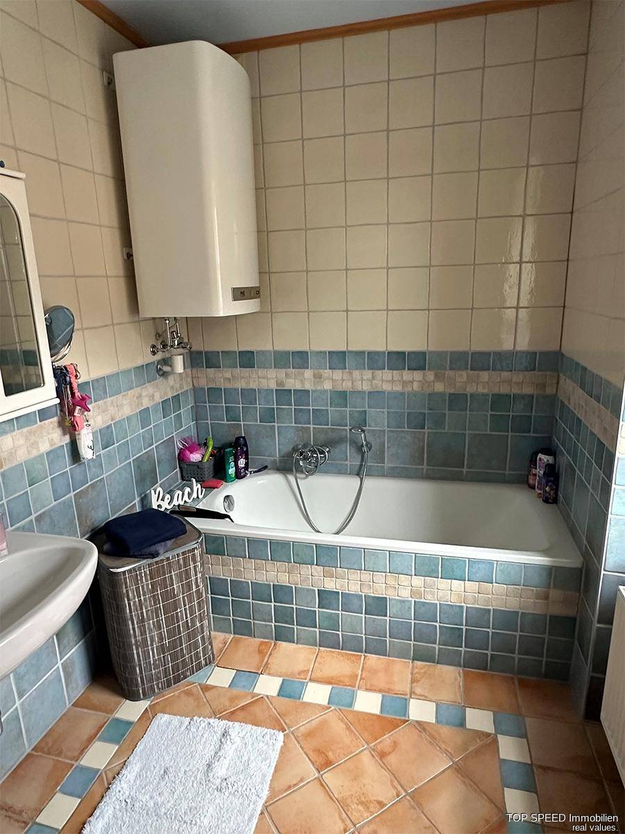Badezimmer mit Badewanne, blau-beigen Fliesen und einem Wandboiler für Warmwasser.
