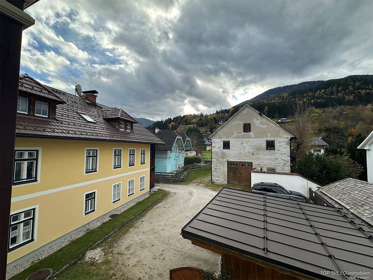 Blick auf umliegende traditionelle Gebäude, eine unbefestigte Straße und bewaldete Berge.