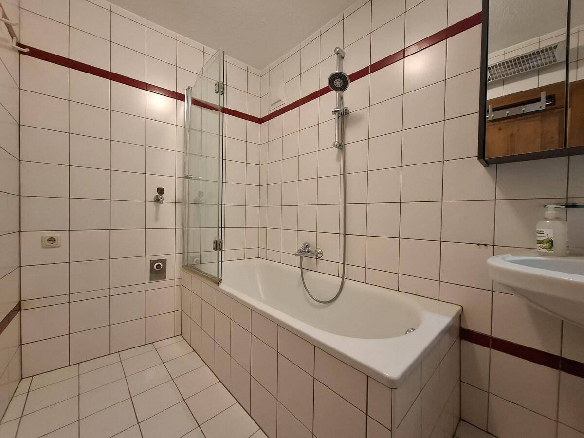 Badezimmer mit weißen Fliesen, Badewanne mit Duschwand und kleinem Waschbecken.