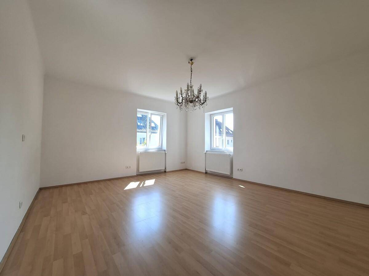 Großes, helles Zimmer mit Parkettboden, Kronleuchter und zwei Fenstern.