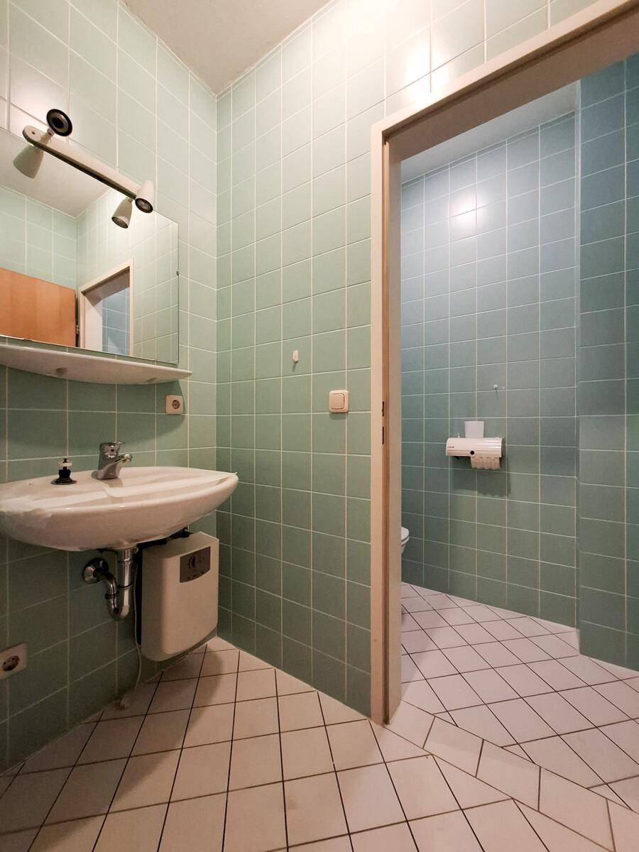 Badezimmer mit hellgrünen Wandfliesen, weißem Fliesenboden, Waschbecken und Spiegel.