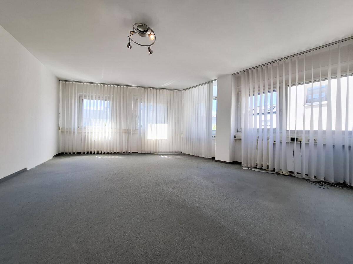 Heller Büroraum mit grauem Teppichboden und vertikalen Jalousien an den Fenstern.