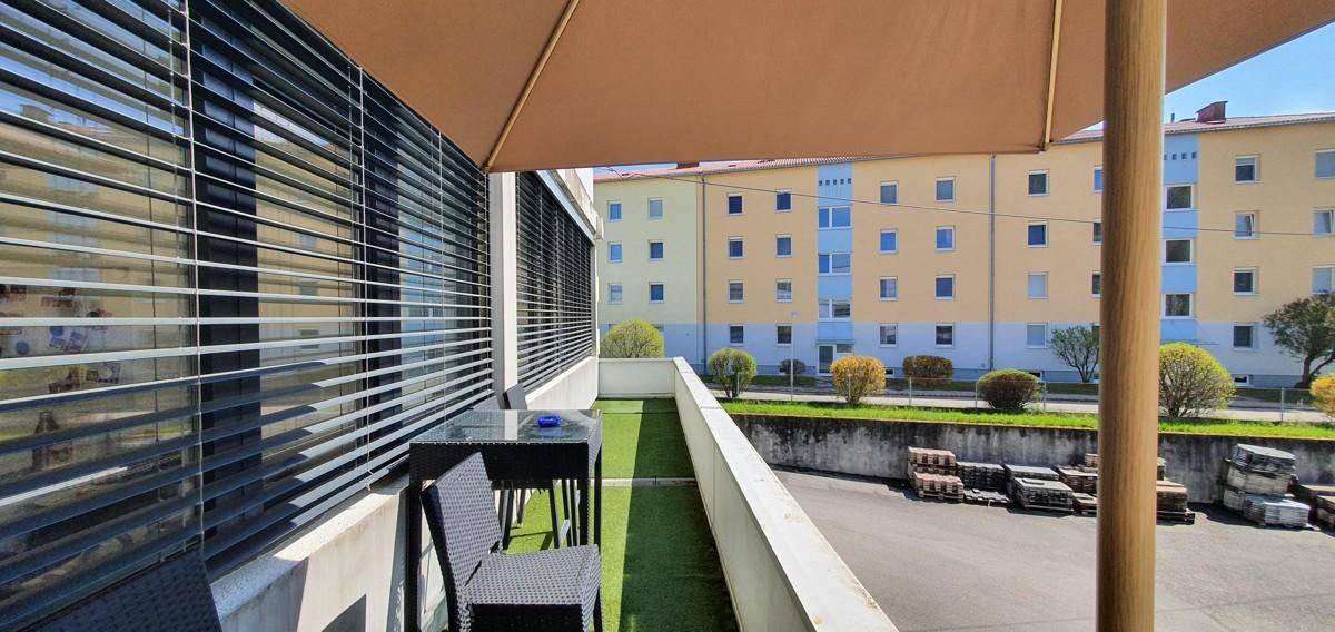 Terrasse mit Kunstrasen, Gartenmöbeln und Blick auf umliegende Wohngebäude unter einem Sonnenschirm.