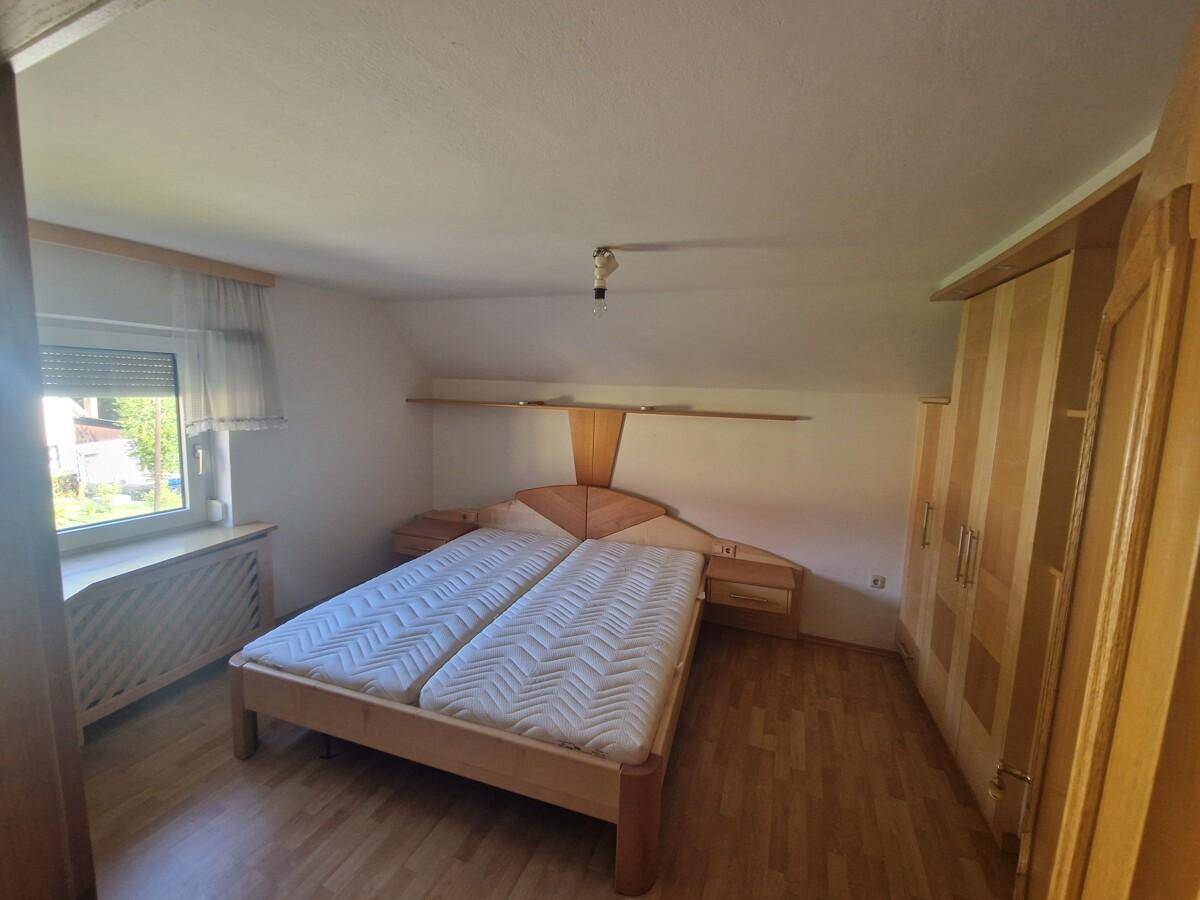 Schlafzimmer mit Doppelbett, Holzschrank und Fenster, das Tageslicht in den Raum lässt.