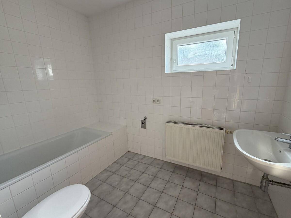 Helles Badezimmer mit Badewanne, Waschbecken und Fenster, ausgestattet mit weißen Fliesen.