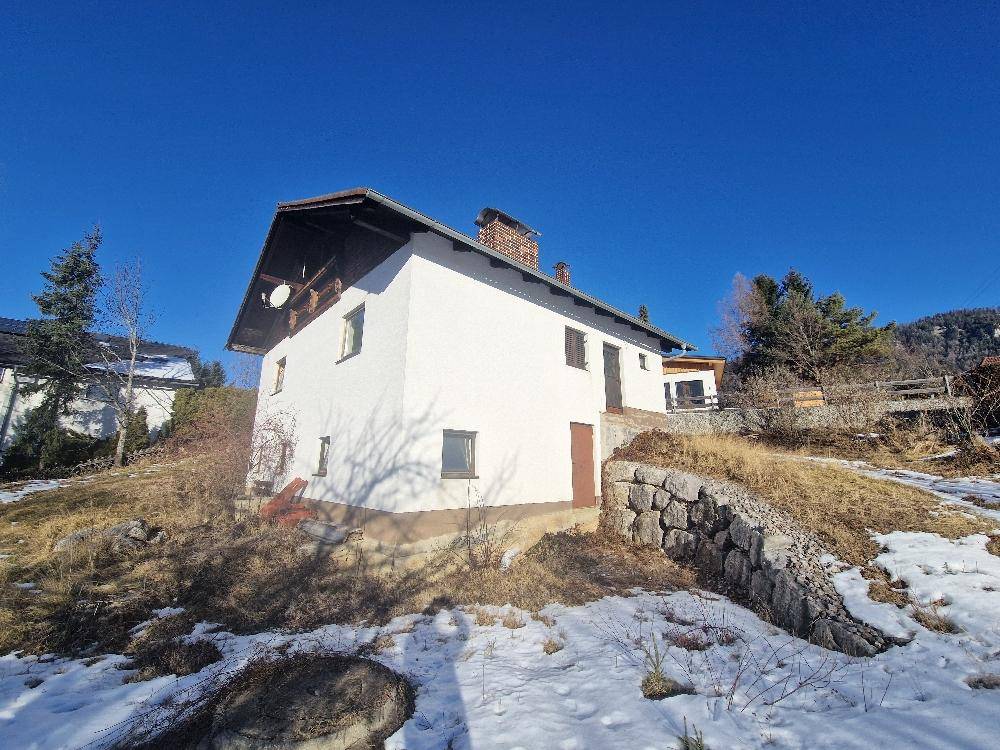 Rückansicht des Hauses mit weißer Fassade und Schornstein, umgeben von Schnee und trockener Vegetation.